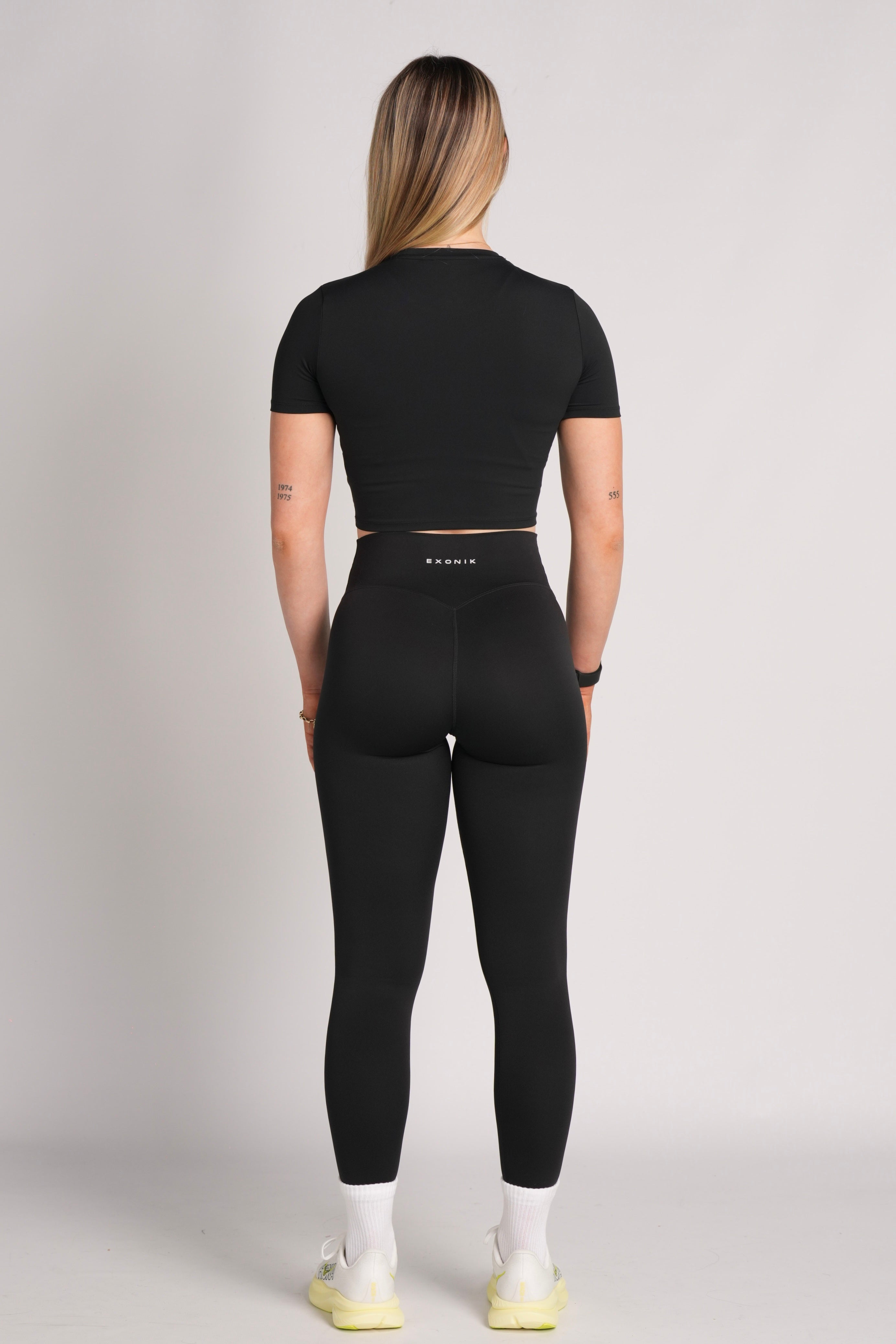 Full-body back view of a model wearing the Exonik ExoTech Liberty Crop Tee in Black in a studio setting.
