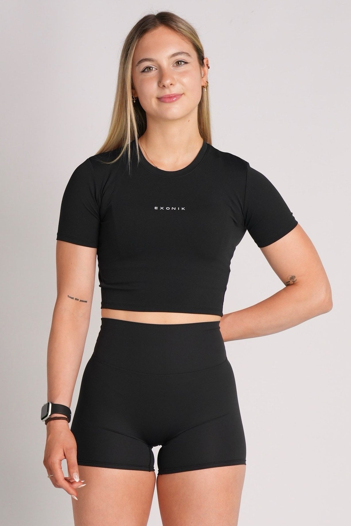 Model in the Exonik ExoTech Liberty Crop Tee in Black, smiling and standing in a relaxed pose against a plain studio backdrop.